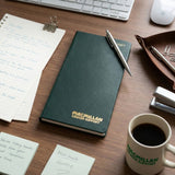 A dark green leather-bound notebook with gold foil 'MACMILLAN CANCER SUPPORT' branding sits on a wooden desk, styled with a silver pen, a coffee mug, sticky notes, and office supplies.